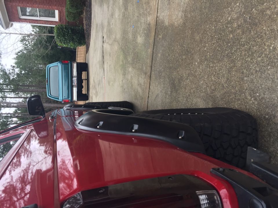 Red vehicle side view with fender flare and knobby tire, teal SUV in background.