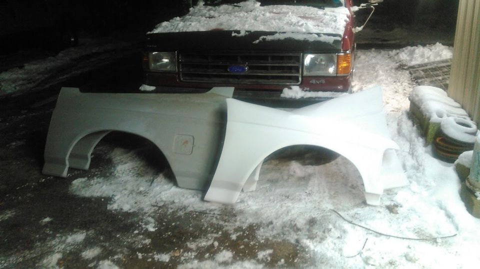 Two primered front fenders in snow in front of a dark red Ford 4x4 truck at night