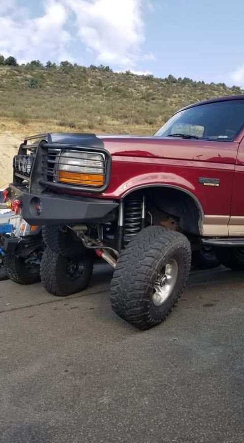1996 Ford Bronco - The front quarter panel and custom bumper of a lifted, dark red Ford Bronco are visible. The vehicle is parked on asphalt with a dry, grass…