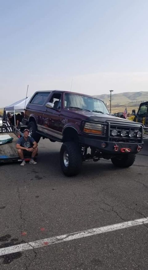 1996 Ford Bronco - A man crouches on a trailer next to a lifted, dark red 1996 Ford Bronco with a custom front bumper and auxiliary lights. The vehicle is par…