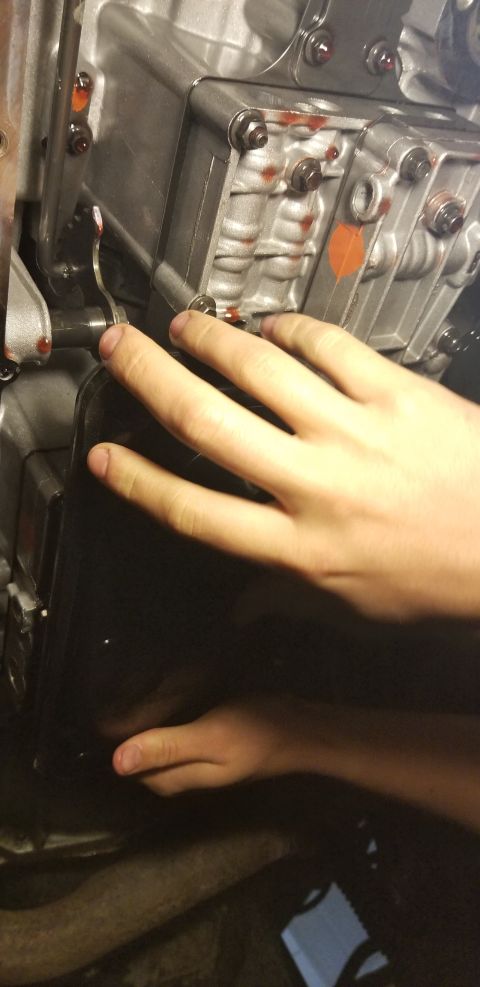 1996 Ford Bronco - A person's hands are shown reaching towards a dark, rectangular transmission pan on a vehicle. Red fluid is visible on the metal casing abo…