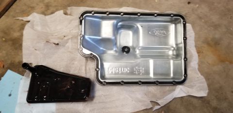 1996 Ford Bronco - A new, shiny Ford transmission pan with a gasket is laid out on white paper next to a used, dark transmission filter. The pan has a Ford lo…