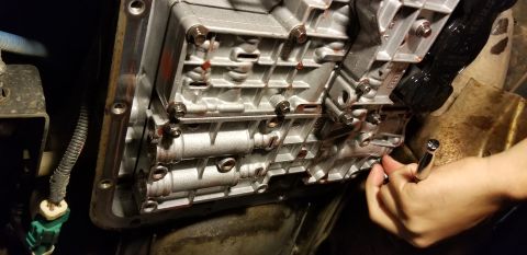 1996 Ford Bronco - A mechanic's hand uses a socket wrench to tighten a bolt on the transmission pan of a vehicle. The transmission pan is silver and has red f…