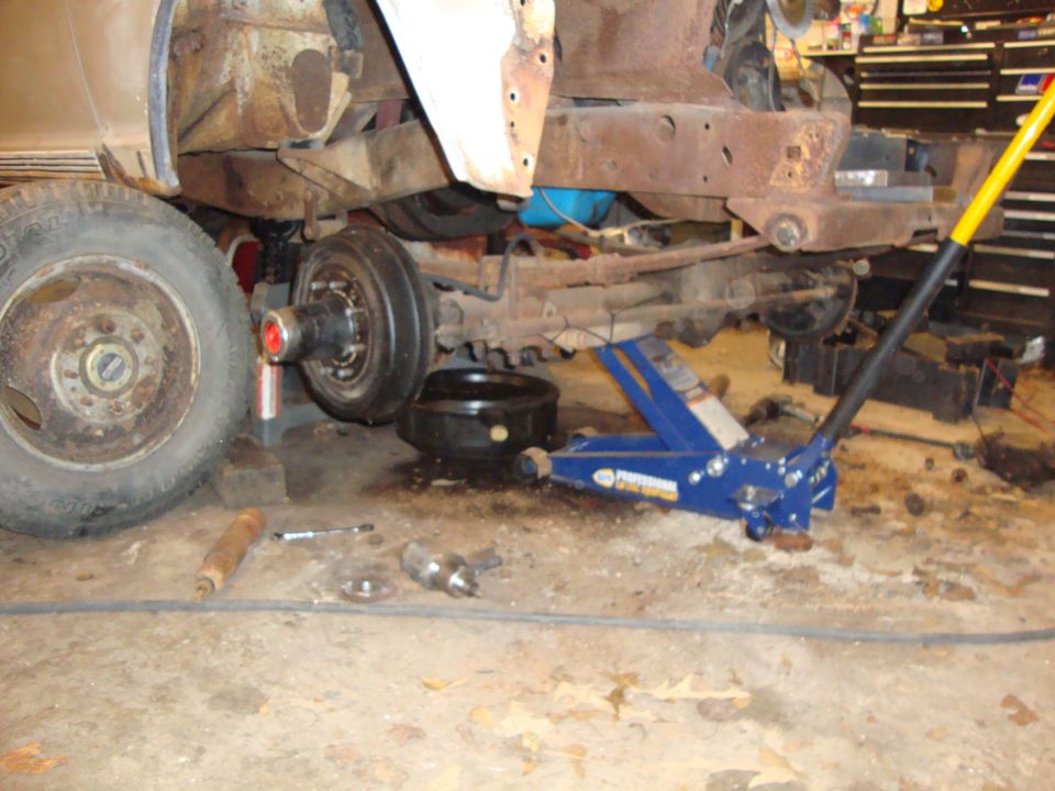 Blue floor jack under a rusty vintage truck frame, front wheel and brake drum visible.