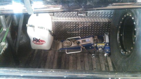 1978 Ford Bronco - The bed of a pickup truck contains a white plastic fluid tank with 'RK Racing' logo, a blue hydraulic jack, and a spare tire. A diamond pla…