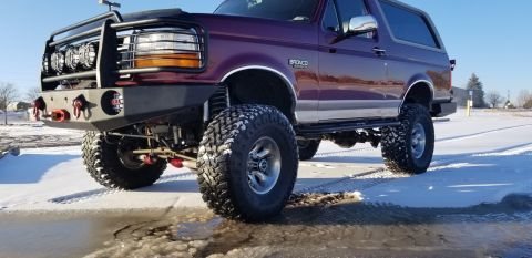 1996 Ford Bronco - A lifted, dark red 1996 Ford Bronco is parked on a snowy and icy surface under a clear blue sky. The vehicle features a custom front bumper…