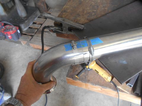 1992 Ford Bronco - A hand holds a curved stainless steel pipe with blue tape and hose clamps attached. A yellow angle grinder rests on a wooden surface nearby…