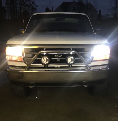 1995 Ford Bronco - The front of a white Ford Bronco is illuminated by its headlights and two KC auxiliary lights mounted on a chrome bull bar. The vehicle is…