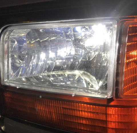 1995 Ford Bronco - A close-up view of the driver's side headlight and amber turn signal of a vehicle. The headlight is illuminated and shows a clear lens with…