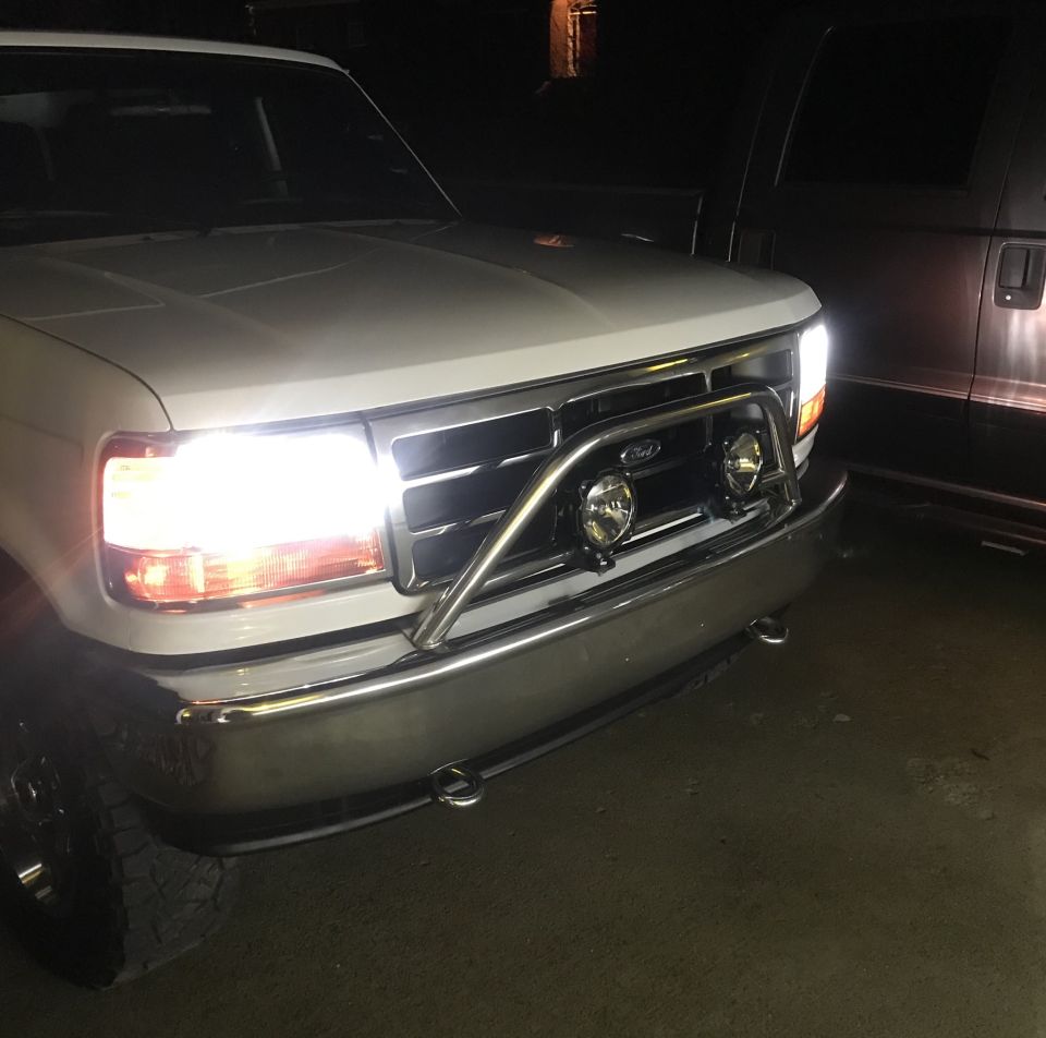 White Ford truck front with chrome bull bar and auxiliary lights, headlights on.