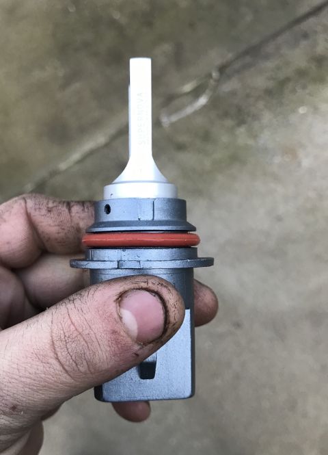 1995 Ford Bronco - A person's dirty hand holds a silver LED headlight bulb with a red O-ring seal. The bulb has "SUPERNOVA" printed vertically on its white up…