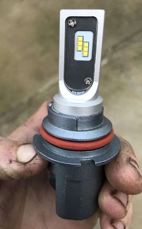 1995 Ford Bronco - A close-up shot shows a person's dirty hand holding a silver and black LED headlight bulb with yellow LEDs visible. The bulb has a gray bas…