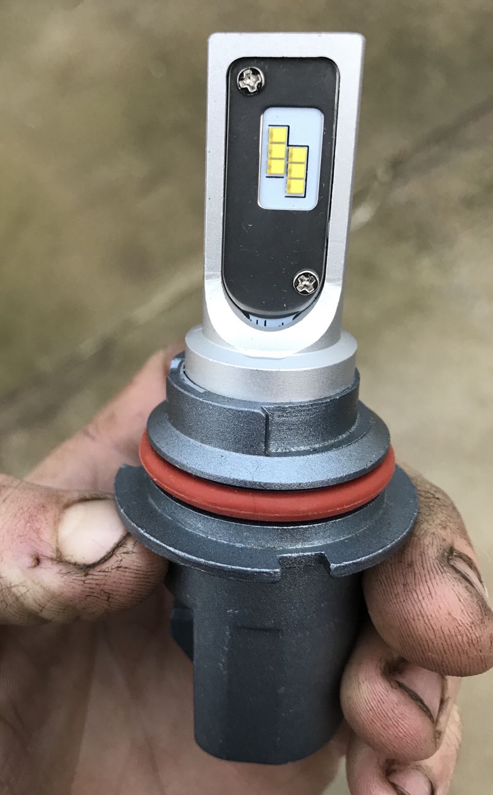 Close-up of a silver and black LED headlight bulb with yellow LEDs, held by a dirty hand.