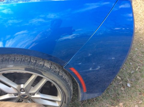 1992 Ford Bronco - A close-up view of the rear quarter panel and tire of a blue vehicle. The body shows some scuff marks and reflections of the sky and clouds.