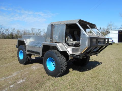 1992 Ford Bronco - A custom-built, all-metal, 4x4 vehicle with large off-road tires and bright blue rims sits parked on a grassy field under a clear blue sky.