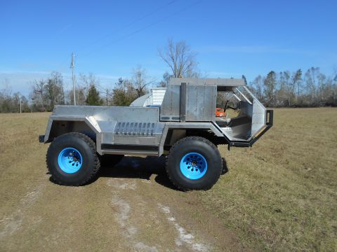 1992 Ford Bronco - A custom-built, open-top, four-wheel-drive vehicle with a brushed aluminum body and bright blue wheels is parked on a dirt path in a grassy…