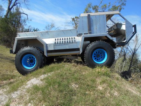 1992 Ford Bronco - A custom-built, silver, off-road vehicle with large tires and bright blue rims sits on a grassy incline under a blue sky.