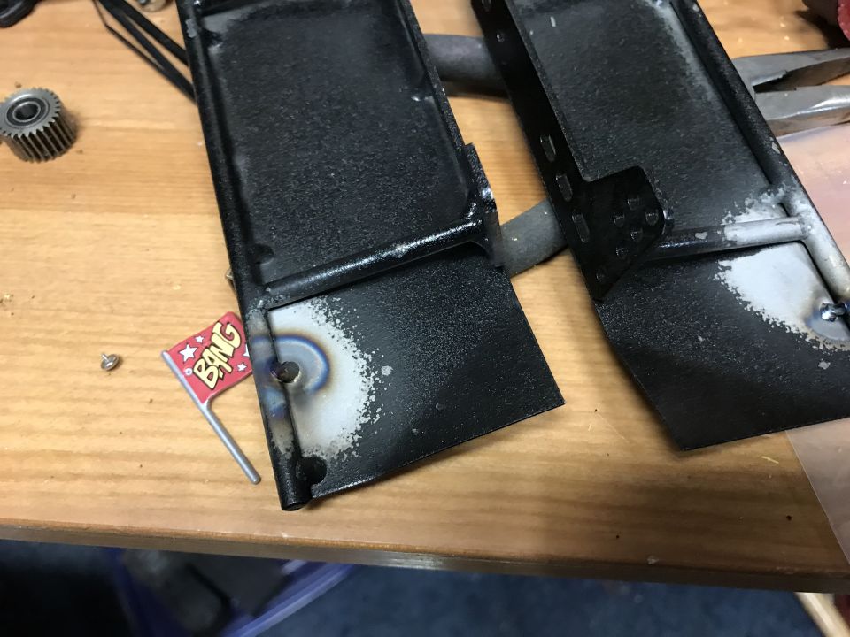 Two black metal parts with welding marks and a small red flag with the word BANG.