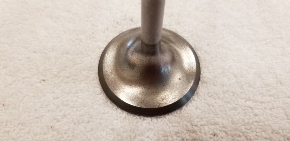 Metal engine valve head and stem on a white textured surface.