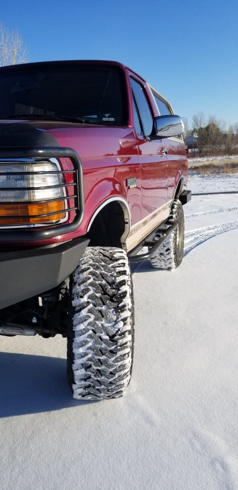 1996 Ford Bronco - A close-up, low-angle shot of the front passenger side of a lifted, maroon 1996 Ford Bronco with aggressive tires covered in snow, parked i…