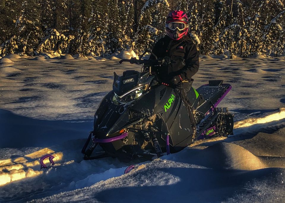 Person on a black Arctic Cat snowmobile with green and purple accents in a snowy forest.
