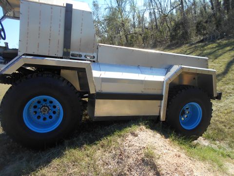 1992 Ford Bronco - A custom-built vehicle with a silver, angular body and large, knobby tires with bright blue rims is parked on a grassy incline. The vehicle…