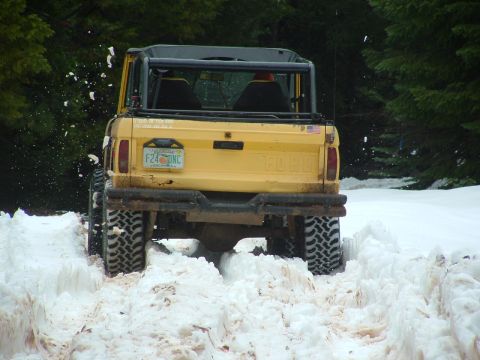 1990 Ford Bronco - Yellow Ford Bronco navigates a snowy trail, kicking up snow with its aggressive off-road tires.