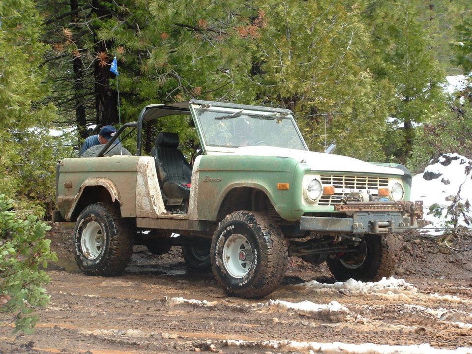 Green Ford Bronco with white top and roll cage on muddy trail, front quarter view