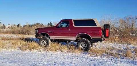 1996 Ford Bronco - A lifted, dark red 1996 Ford Bronco is parked on a snowy, grassy field under a clear blue sky. The SUV features a tan roof, large off-road…