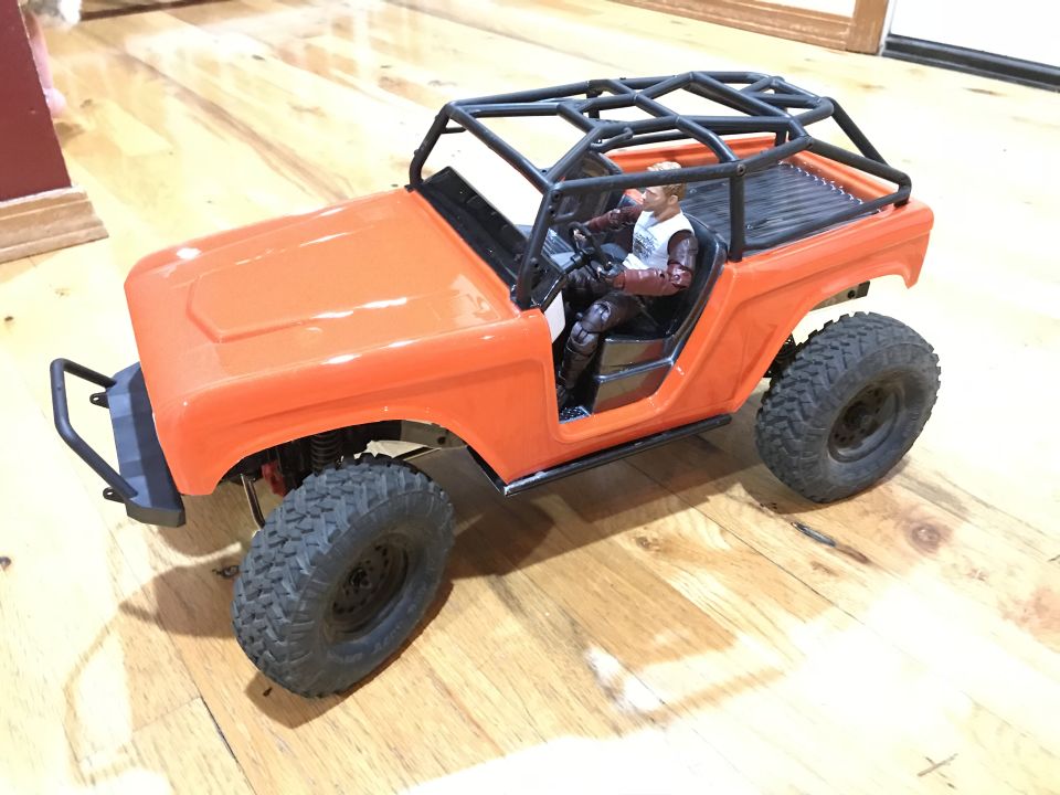 Orange RC rock crawler with driver figure and roll cage on a wooden floor.