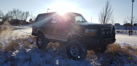 1996 Ford Bronco - A dark-colored Ford Bronco is parked on a snow-covered embankment, with the sun flaring brightly in the upper left corner of the image. The…
