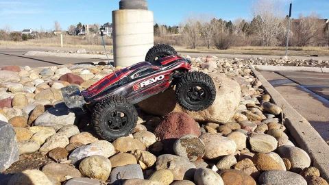 1996 Ford Bronco - A red and black remote-controlled monster truck with off-road tires is positioned on a pile of large river rocks. The truck has the word "R…
