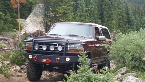 1996 Ford Bronco - A dark red 1996 Ford Bronco is shown driving on a rocky, wooded trail. The vehicle has a black bull bar with four auxiliary lights and a re…