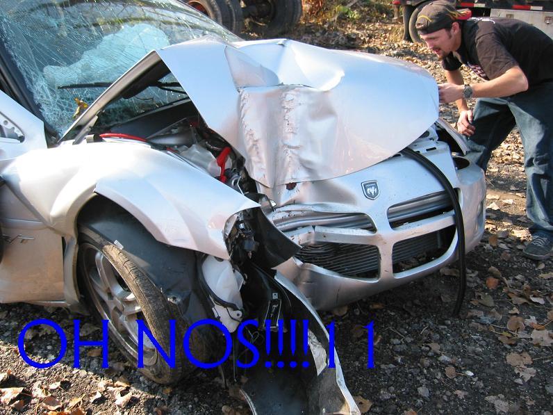 Silver car with crumpled hood, shattered windshield, and damaged front bumper. A man inspects the damage