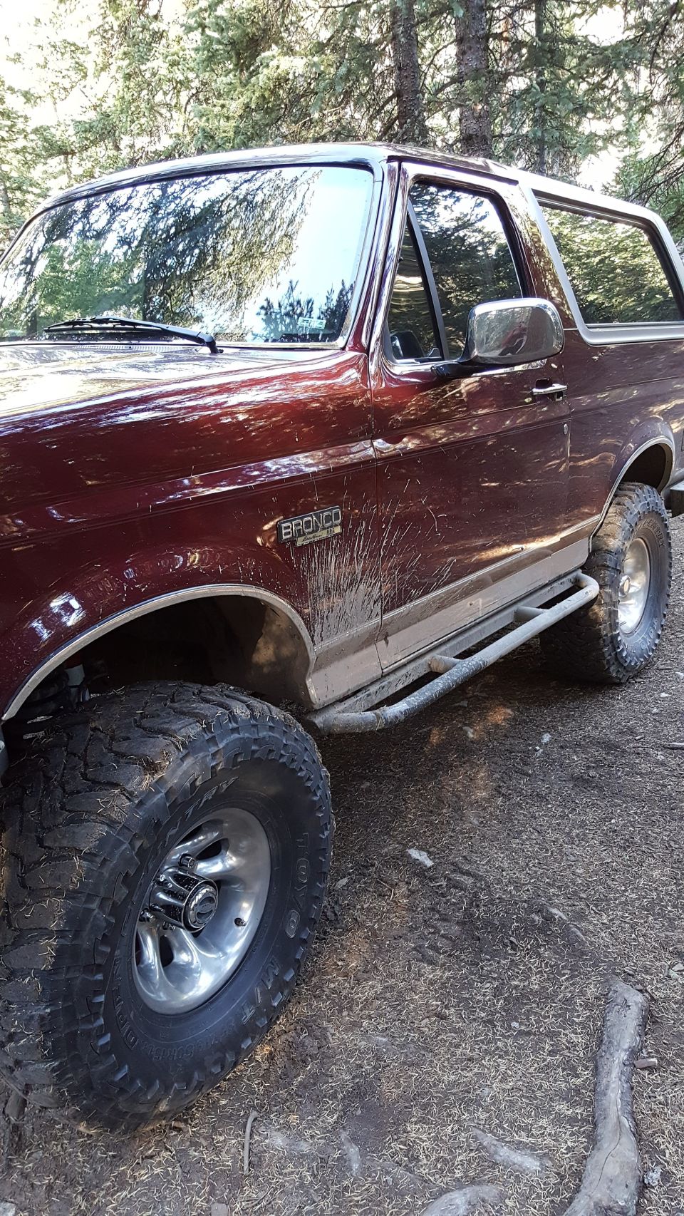 Maroon Ford Bronco with mud splashes, off-road tires, and side steps parked in a wooded area.