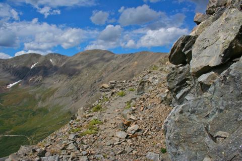 1996 Ford Bronco - A rocky mountain trail winds along a steep slope under a blue sky with scattered clouds. In the distance, rugged mountain peaks rise agains…