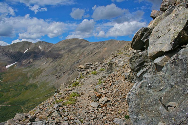 Rocky mountain trail with distant peaks under a cloudy blue sky.