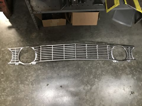 1964 Ford Fairlane - A chrome front grille with circular headlight cutouts is laid flat on a concrete floor. The grille has horizontal bars and a central peak.