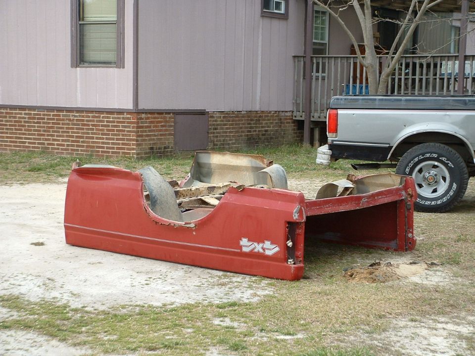 Red Ford truck bed shell removed and sitting on the ground in a yard, gray pickup parked behind