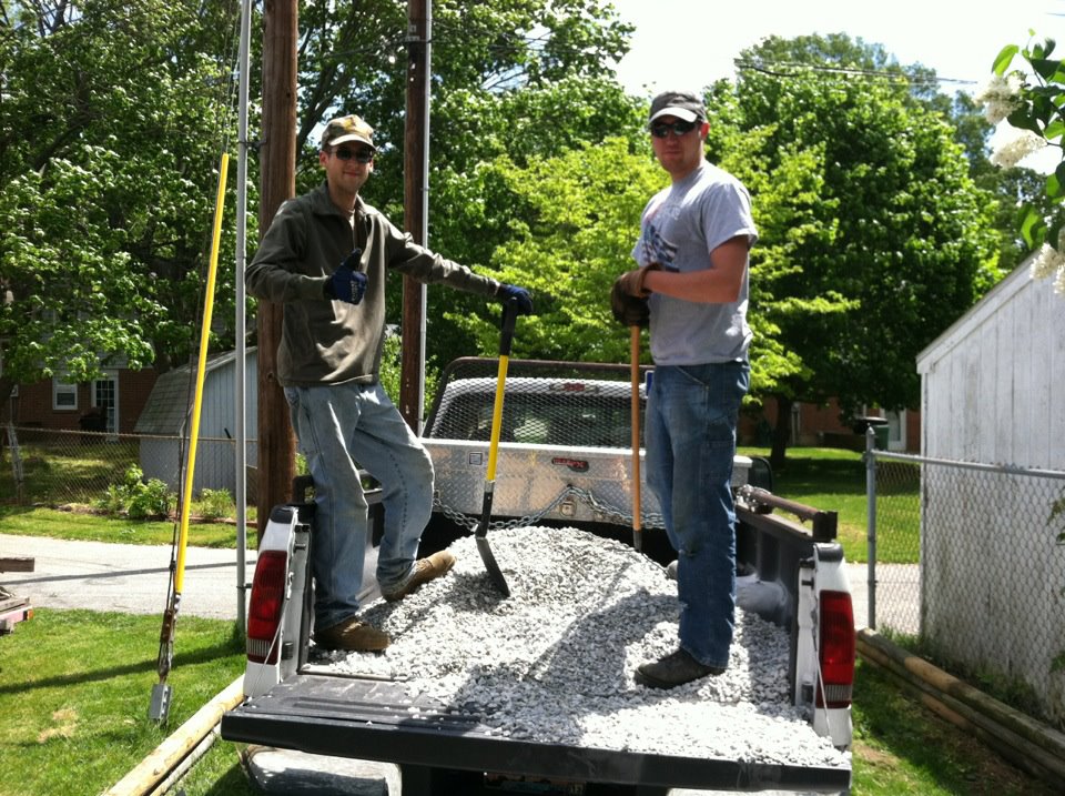 Two men stand in the bed of a pickup truck loaded with gravel, holding shovels