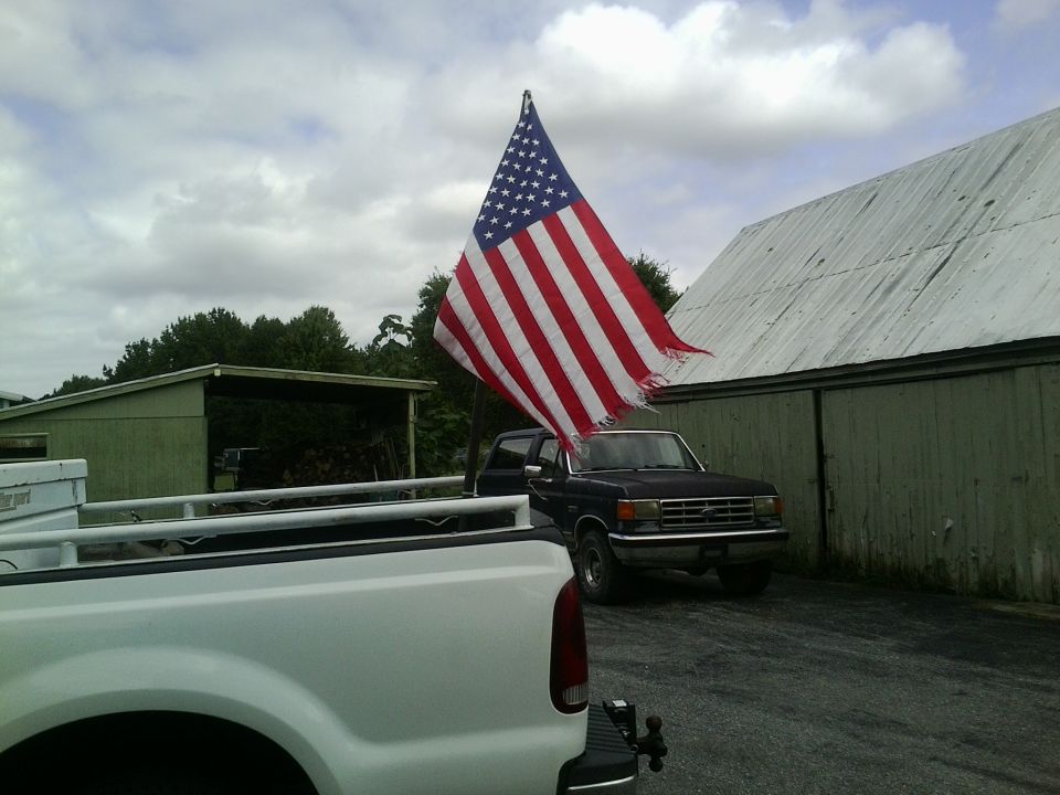 Dark blue Ford F-Series truck with American flag flying, parked near a barn. White truck in foreground