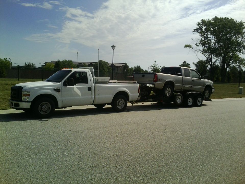 White Ford F-250 towing a silver Chevrolet Silverado 2500HD on a triple-axle trailer