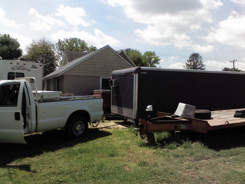 White pickup truck with open driver door, towing a black enclosed trailer and a rusty flatbed trailer in a grassy yard