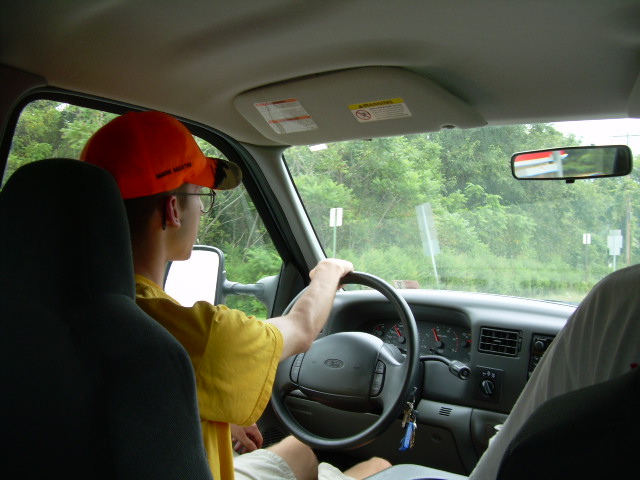 Driver wearing an orange hat and glasses, hands on the steering wheel of a truck. Dashboard and trees visible outside