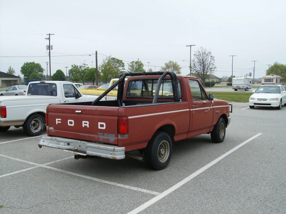 Red 1987 Ford F-150 pickup truck with a black roll bar in the bed, parked in a lot next to a white Ford pickup