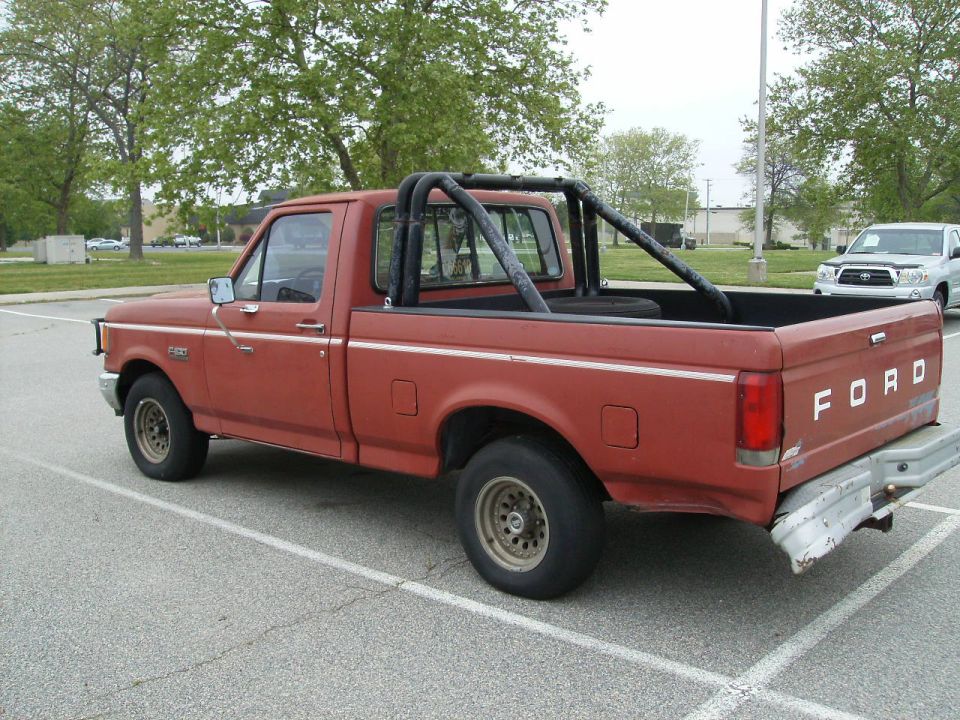 Red 1987 Ford F-150 pickup truck with roll bar and spare tire in bed, parked in lot. White SUV in background