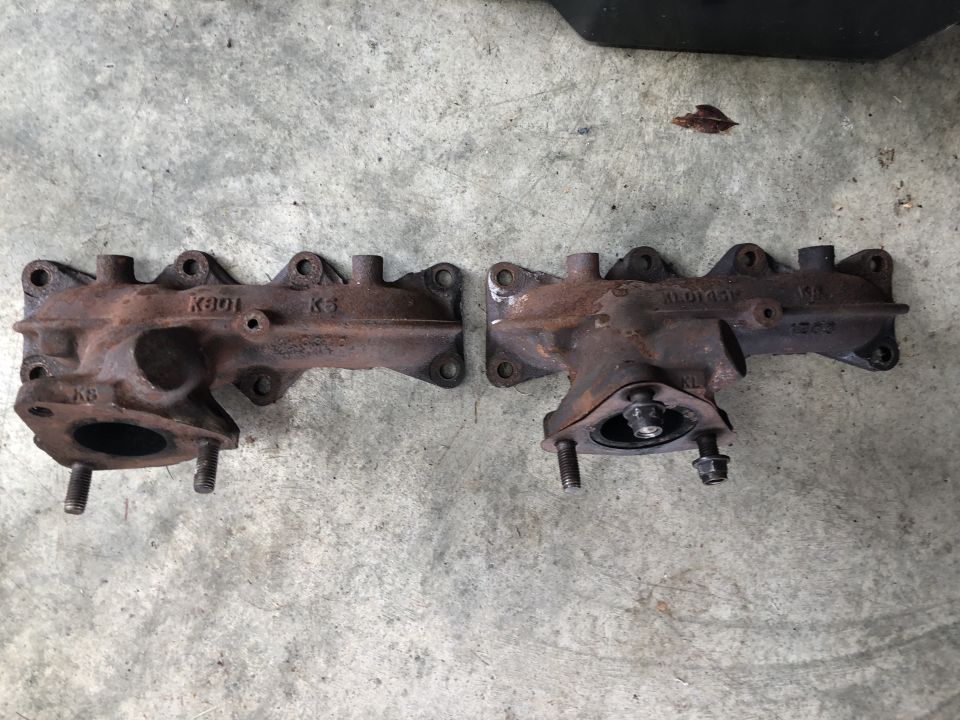 Two rusty exhaust manifolds on a concrete surface. Text visible on manifolds