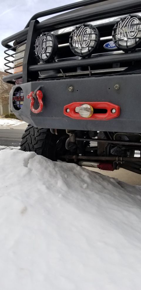 1996 Ford Bronco - A close-up, low-angle shot of the front bumper and grille area of a modified vehicle, featuring KC lights, a red tow hook, and a red winch…