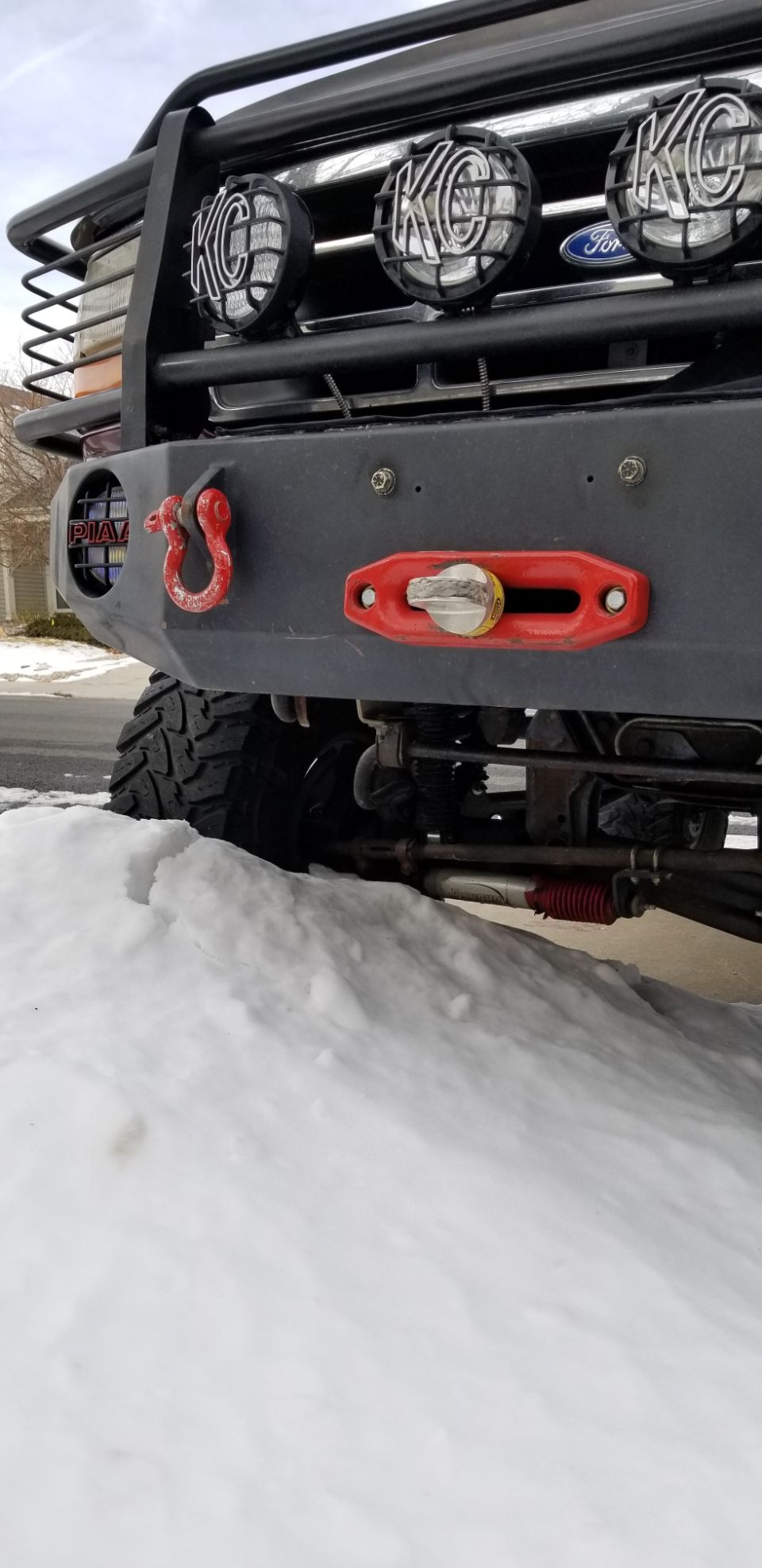 Front bumper of a modified vehicle with KC lights, red tow hook, and winch fairlead, partially covered in snow.