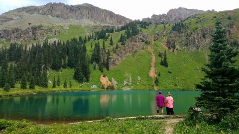1996 Ford Bronco - Two people stand at the edge of a clear turquoise lake, looking out at the water with a steep, grassy mountainside and rocky peaks in the b…